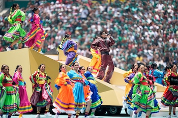 Un cantante con chaqueta de cuero oscuro y gafas de sol en una plataforma dorada, rodeado de muchos bailarines folclóricos con trajes brillantes y coloridos