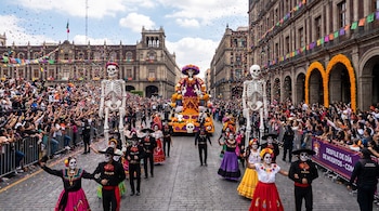 Desfile con personas disfrazadas de catrinas y calaveras, esqueletos gigantes, carrozas alegóricas, público, edificios decorados con papel picado y flores.