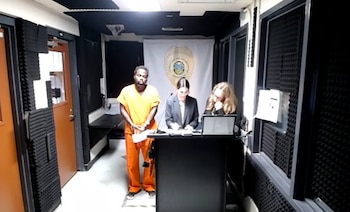 Un hombre con uniforme naranja de prisión permanece de pie junto a dos mujeres detrás de un podio en una sala