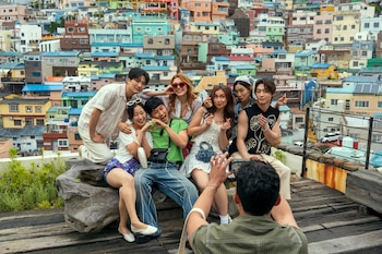 Un grupo de siete jóvenes, incluyendo Anna Cathcart y Gia Kim, posa sonriendo frente a un pueblo coreano de casas coloridas, mientras un octavo toma una foto