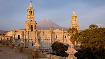 Vista de la Catedral de Arequipa, de piedra blanca con dos torres, bajo un cielo azul claro. Detrás, el volcán Misti con su cima nevada. Árboles verdes a la derecha
