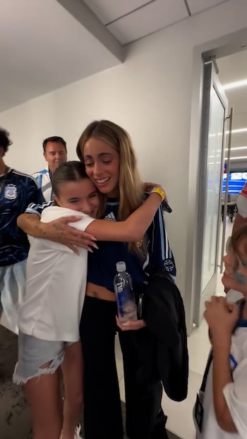 Mujer rubia, Tini Stoessel, con camiseta deportiva azul y blanca, abraza a una niña con camiseta blanca. Se observa un hombre en segundo plano y una puerta