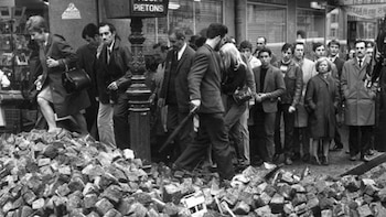 Fotografía en blanco y negro de personas sorteando una gran barricada de adoquines en una calle urbana; edificios y un poste de luz son visibles