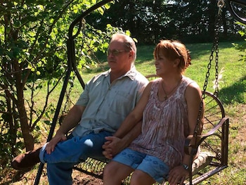 Hombre y mujer sentados en un columpio de jardín, tomados de la mano. Él usa camisa clara, jeans y botas; ella, blusa sin mangas y shorts. Fondo de árboles y césped