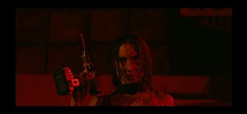 Magui Bravi, con el rostro sucio y sangre, sostiene un taladro mientras mira fijamente a cámara, bajo una intensa luz roja que ilumina la escena