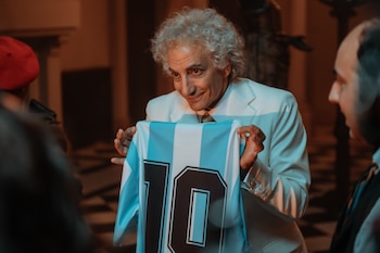 Un hombre con traje blanco y cabello rizado canoso sostiene una camiseta de fútbol a rayas celestes y blancas con el número diez