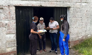 Tres mujeres con mascarillas y carpeta de apuntes entregan un paquete a un hombre mayor en la entrada de una casa de piedra