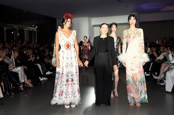 Modelos con vestidos florales y una mujer con traje negro caminan por una pasarela interior rodeada de espectadores