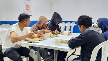 Varias personas sentadas en una mesa blanca comiendo de bandejas de comida en un comedor social, con sillas blancas y una pared azul al fondo
