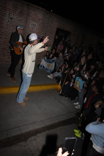 Un cantante con gorra y suéter blanco se dirige a un grupo de personas que lo fotografían de noche frente a un muro de ladrillos