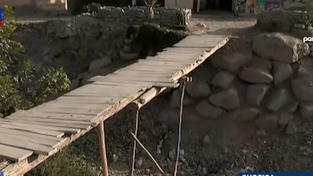 Un puente de tablas de madera sostenido por postes cruza una zanja de tierra y piedras entre dos bordes