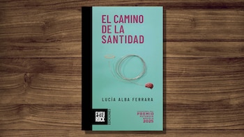 Libro con portada azul claro y texto rojo "El camino de la santidad", firmado por Lucía Alba Ferrara, un halo, un cable en espiral y un cerebro. Se apoya sobre madera