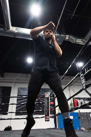 Un hombre con camiseta oscura y pantalones cortos entrena dentro de un ring de boxeo. Se ven cuerdas, focos en el techo y conductos de ventilación