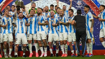 Jugadores de la Selección Argentina, incluyendo a Lionel Messi con el trofeo, celebran en el campo. Visten camisetas celestes y blancas, con confeti cayendo
