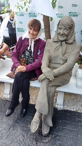 Mujer mayor con pañuelo y chaqueta morada sentada en banco blanco junto a estatua de mujer con pañuelo. Fondo blanco con logos de SITRAM y hojas