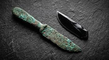 Bisturí de bronce poroso con óxido verde junto a una navaja de obsidiana negra sobre una superficie de piedra oscura.