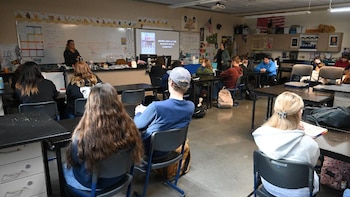 Un aula vista desde atrás, con estudiantes sentados en sillas de oficina en mesas negras, y una profesora de pie al frente junto a una pantalla con texto