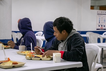 Vista de perfil de un hombre comiendo de una bandeja en un comedor social, con otras personas y mesas blancas al fondo