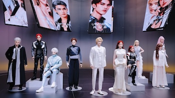 Nueve robots humanoides con vestuario de moda, de pie y sentados en podios, frente a varias pantallas verticales con imágenes de personajes similares