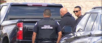 Un agente de espaldas con un chaleco que dice negociador policial y dos hombres de pie entre dos vehículos