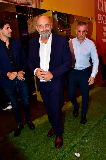Tres hombres caminan sobre una alfombra verde; el del centro, calvo y con barba, viste traje azul oscuro y camisa blanca, mirando al frente