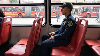 Un agente de la Policía de Colombia con uniforme azul y gorra está sentado en un asiento rojo de TransMilenio, mirando por la ventana hacia otro autobús rojo en la calle.