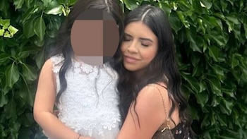 Una mujer de cabello oscuro abraza a una niña con vestido blanco y rostro pixelado frente a un muro de hojas verdes