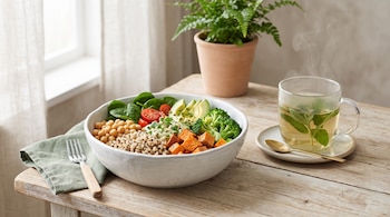 Un plato hondo con garbanzos, quínoa, verduras y aguacate junto a una taza de té humeante sobre una mesa de madera.