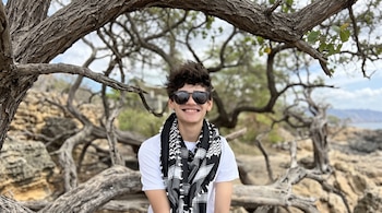 Un joven con gafas de sol, camiseta blanca y pañuelo al cuello sonríe bajo el marco que forman las ramas de un árbol.