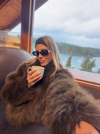 Cande Ruggero con gafas de sol y abrigo de piel sintética sostiene una taza blanca junto a una ventana con vista a un lago