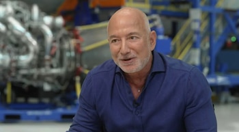 Un hombre calvo con barba gris, Jeff Bezos, lleva una camisa azul oscura y sonríe mientras habla en una entrevista. Un equipo industrial borroso de fondo