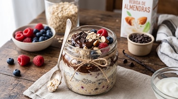 Frasco con avena, chocolate y frutas junto a una cuchara, un tazón con frutos rojos, un bol con yogur y un envase de bebida de almendras.
