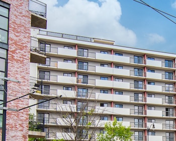 Fachada de un edificio residencial de diez pisos con balcones y ventanas, junto a un edificio de ladrillo bajo un cielo nublado