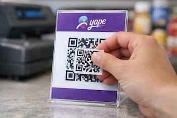 Una mano sostiene una etiqueta adhesiva con código QR sobre un cartel de mostrador transparente que muestra el logo de Yape y otro código QR.