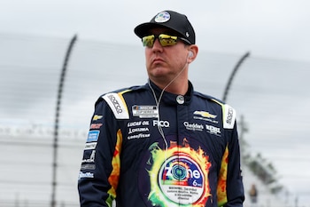 Kyle Busch con un traje de piloto azul marino y amarillo, gorra negra, gafas de sol espejadas y auriculares en un circuito