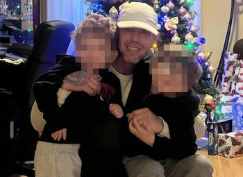 Un hombre con gorra blanca y tatuajes en los brazos abraza a dos niños pequeños, sus caras pixeladas, delante de un árbol de Navidad decorado y regalos