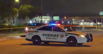 Un patrullero blanco de la policía de Miami-Dade con luces encendidas estacionado de noche tras una cinta de seguridad amarilla