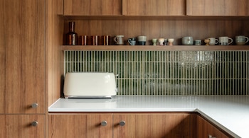 Cocina con armarios de madera y encimera blanca. Tostadora crema en primer plano. Salpicadero de azulejos verdes verticales y estantes con tazas y botellas.