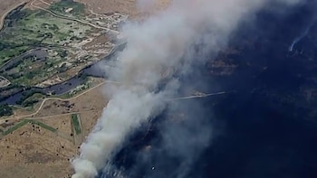 Vista aérea de un paisaje donde una gran columna de humo gris y blanco se eleva sobre una zona industrial o refinería, con campos, carreteras y un río