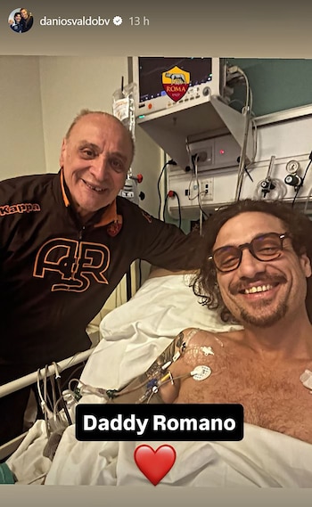 Daniel Osvaldo recostado en una cama de hospital con electrodos en el pecho sonríe junto a un hombre con indumentaria deportiva