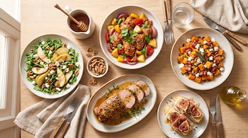 Platos de cerámica sobre una mesa de madera con pollo, lomo de cerdo, ensalada de pera, calabaza asada y tostadas con higos.
