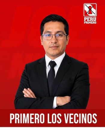Un hombre con traje, anteojos y brazos cruzados posa frente a un fondo rojo con el logotipo del partido Perú Primero