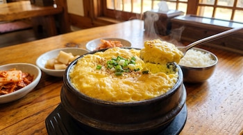 Un cuenco negro con huevo al vapor decorado con cebollín, una cuchara que sostiene una porción, un tazón de arroz y platos con kimchi.