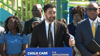 Un hombre con barba y traje oscuro habla en un podio con las palabras "CHILD CARE". Detrás de él, varias personas escuchan en un parque infantil