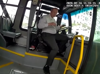Un hombre vestido de chofer de autobús sentado al volante, mirando hacia adelante. Dentro de un autobús con asientos vacíos y ventanas mostrando un edificio