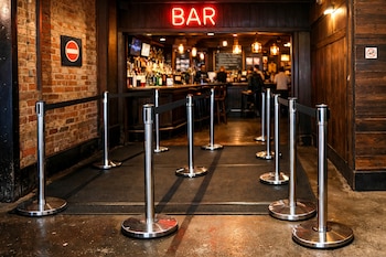 Entrada a un bar con un letrero de neón rojo, postes metálicos con cintas negras para organizar filas, una pared de ladrillo y una pared de madera.