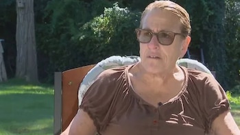 Una mujer con gafas y blusa marrón está sentada en un sillón en un jardín con césped y árboles al fondo