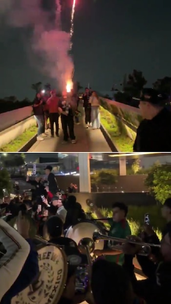 Una multitud de personas en la noche, con fuegos artificiales iluminando el cielo y una banda de música con bombos y trompetas