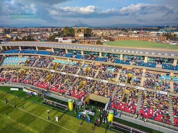 Tribunas con espectadores en un estadio junto a una cancha verde, con una zona urbana e iglesia al fondo