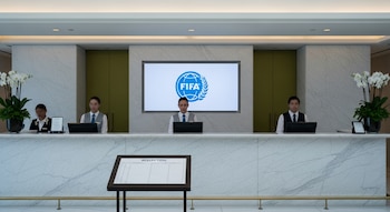 Recepción de hotel con mostrador de mármol claro, tres empleados uniformados, el logo azul de la FIFA en una pantalla digital y un tablero de reservas.